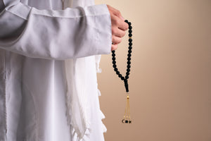 Model holding a gold tasbih with black beads and a gold tassel featuring three pendants, with the central square pendant containing a black nano chip inscribed with the entire Quran, photographed against a beige-toned background.