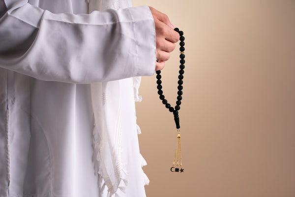 Model holding a gold tasbih with black beads and a gold tassel featuring three pendants, with the central square pendant containing a black nano chip inscribed with the entire Quran, photographed against a beige-toned background.