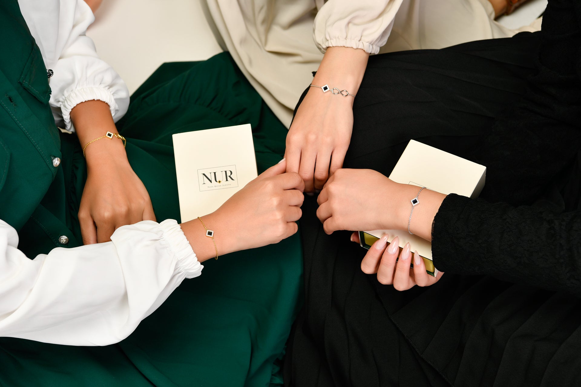 The image shows three women sitting together, each wearing elegant bracelets featuring a small geometric pendant in gold and blue tones. Their outfits—in green, beige, and black—create a refined contrast. Each woman is holding a stylish light-colored jewelry box with the “NUR” logo. The scene conveys a sense of unity, sophistication, and modern luxury, highlighting both the bracelets and the premium packaging.