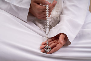 Model holding a silver tasbih with white stones and a silver pendant that includes a Quran-inscribed chip, photographed against a soft beige-toned background.