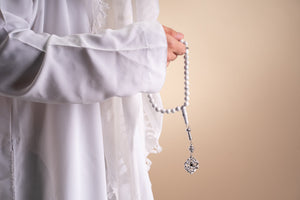 Model holding a silver tasbih with white stones and a silver pendant that includes a Quran-inscribed chip, photographed against a soft beige-toned background.