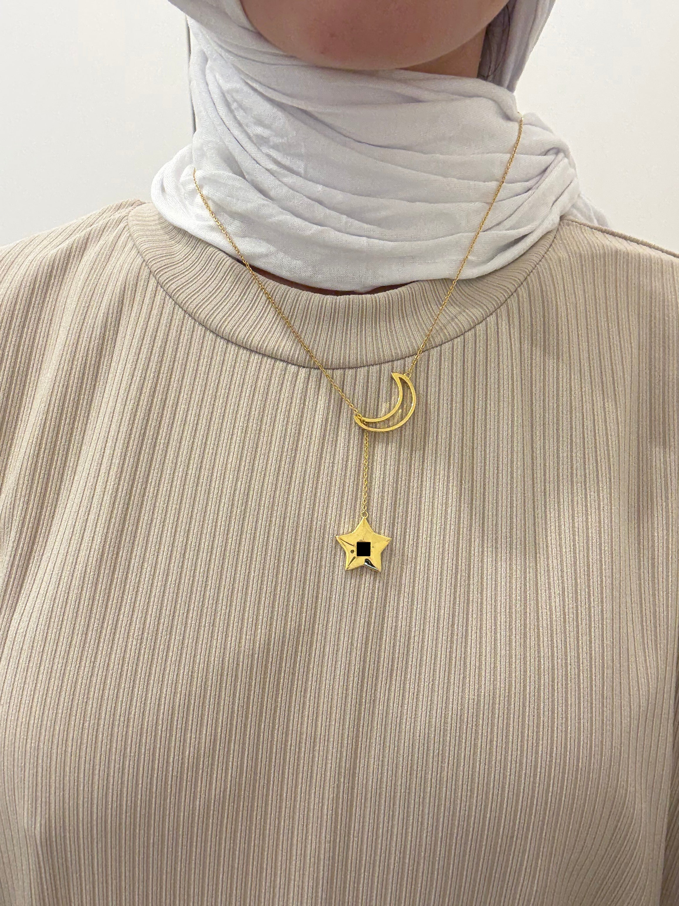 A woman wearing a gold crescent-and-star necklace with the star pendant hanging lower, styled with a beige textured top and a white headscarf, photographed against a soft neutral background.