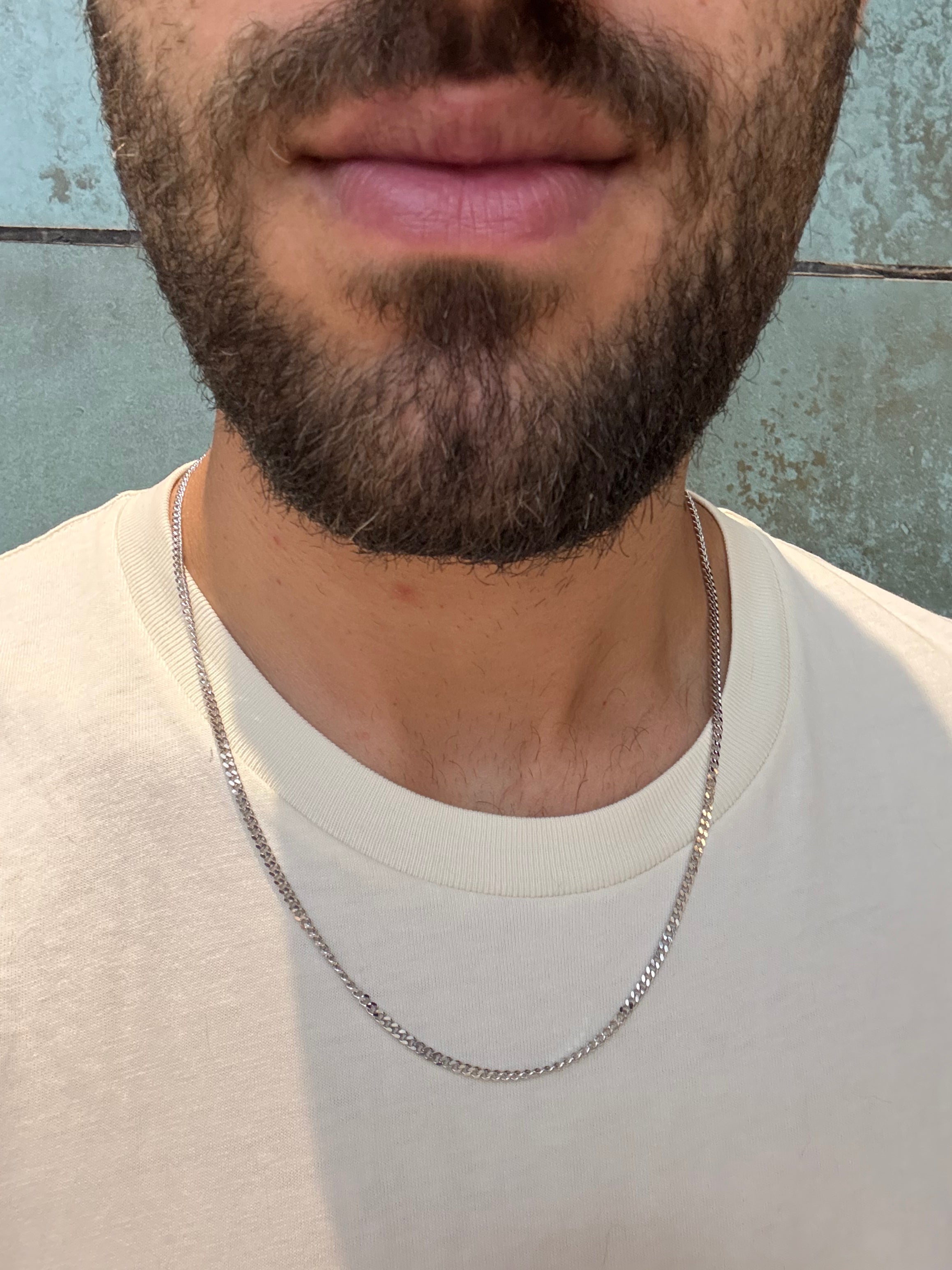 Close-up of a man wearing a silver chain necklace over a light shirt, photographed against a textured wall background.