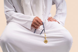 Male model holding a gold tasbih with blue beads and a gold pendant that includes a Quran-inscribed nano chip, photographed against a beige-toned background.