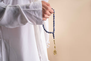 Male model holding a gold tasbih with blue beads and a gold pendant that includes a Quran-inscribed nano chip, photographed against a beige-toned background.