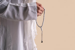 Model holding a silver tasbih with white stones and a silver pendant that includes a Quran-inscribed nano chip, photographed against a soft beige-toned background.