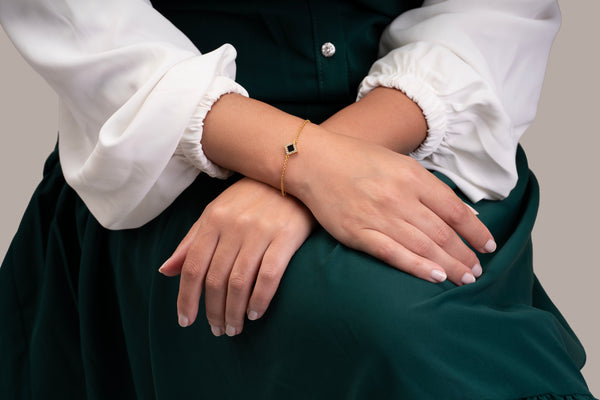 Model wearing a gold bracelet featuring a square black nano-chip charm that contains the full Quran, photographed against a beige-toned background.